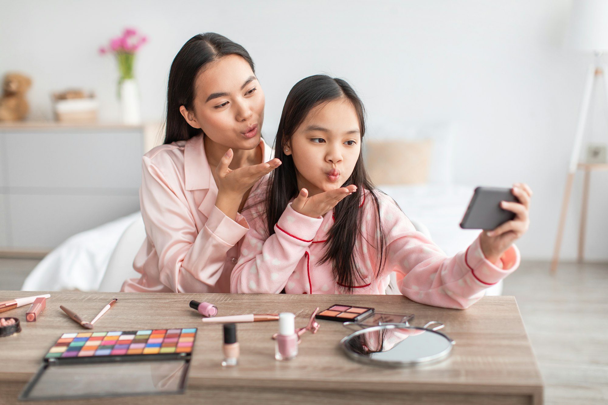 Happy pretty cute asian teen girl and young female in pink pajamas make air kiss and selfie at table