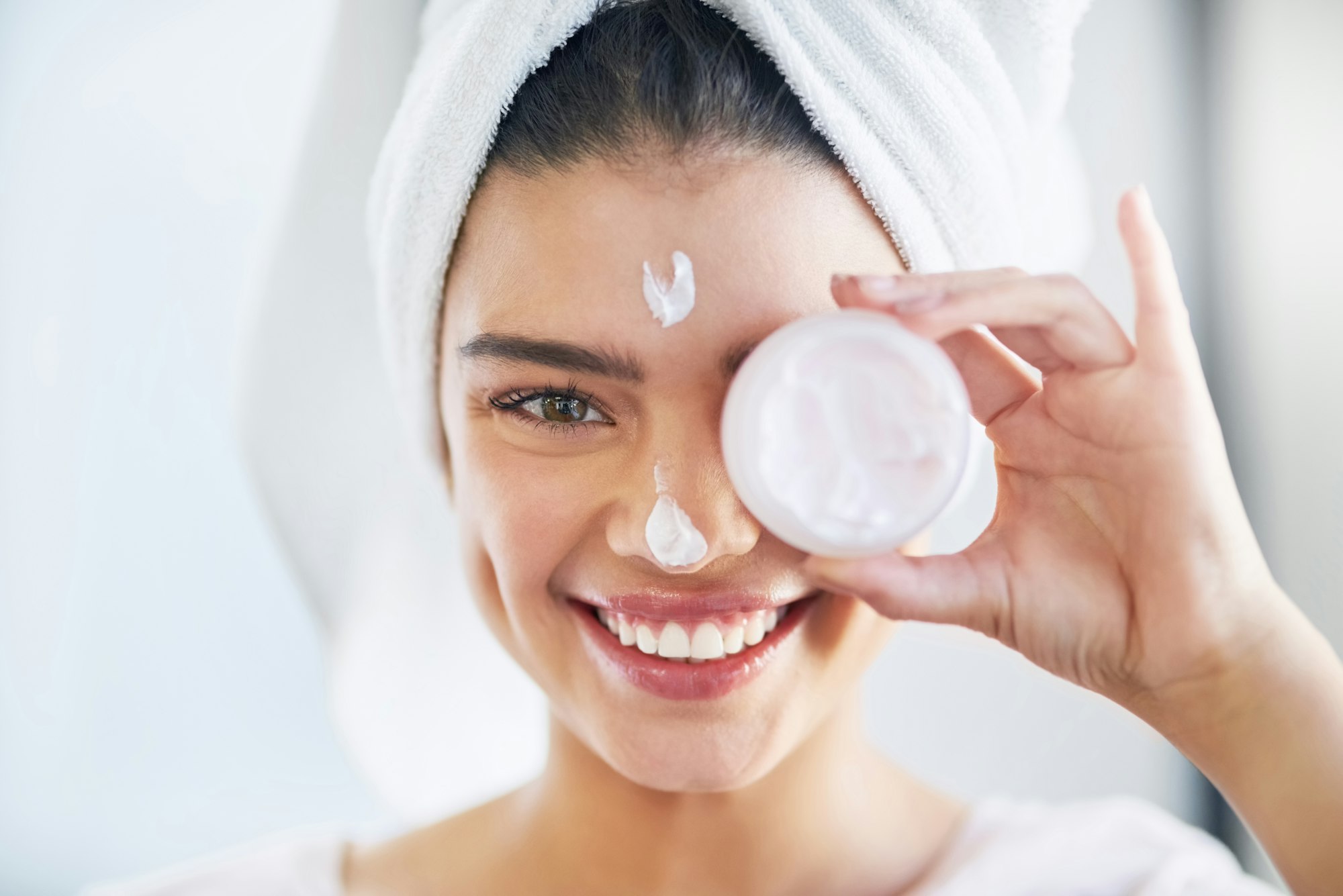 Portrait of a beautiful young woman applying moisturizer to her skin in the bathroom at home